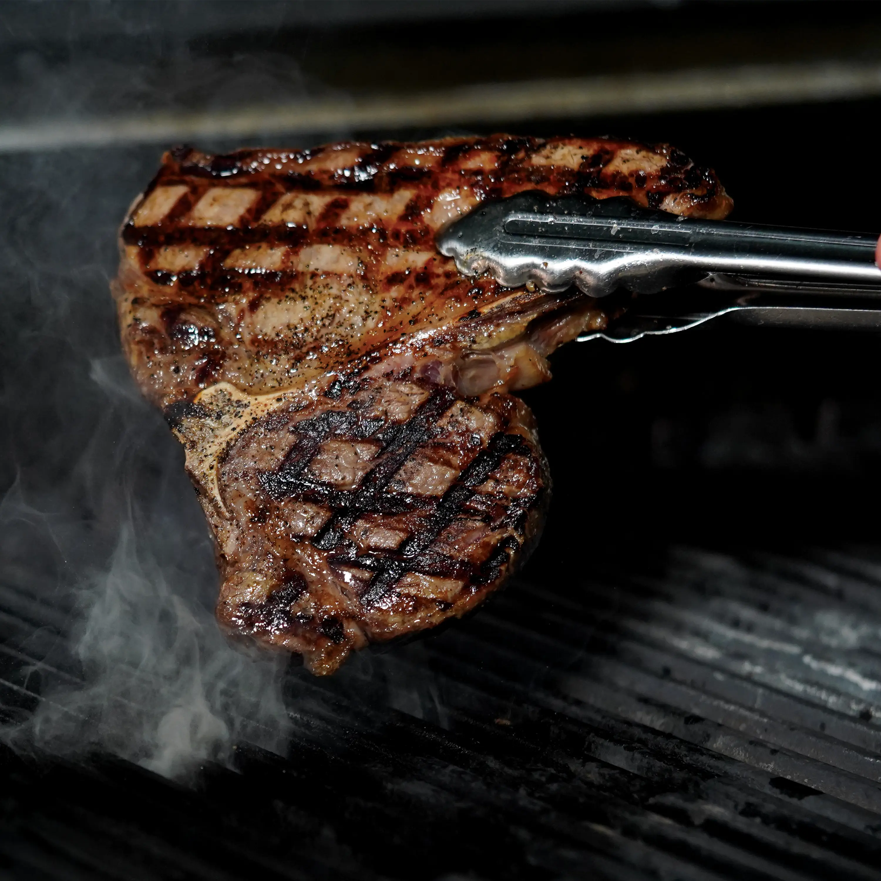 cooked steak held by metal tongs over smoking grill