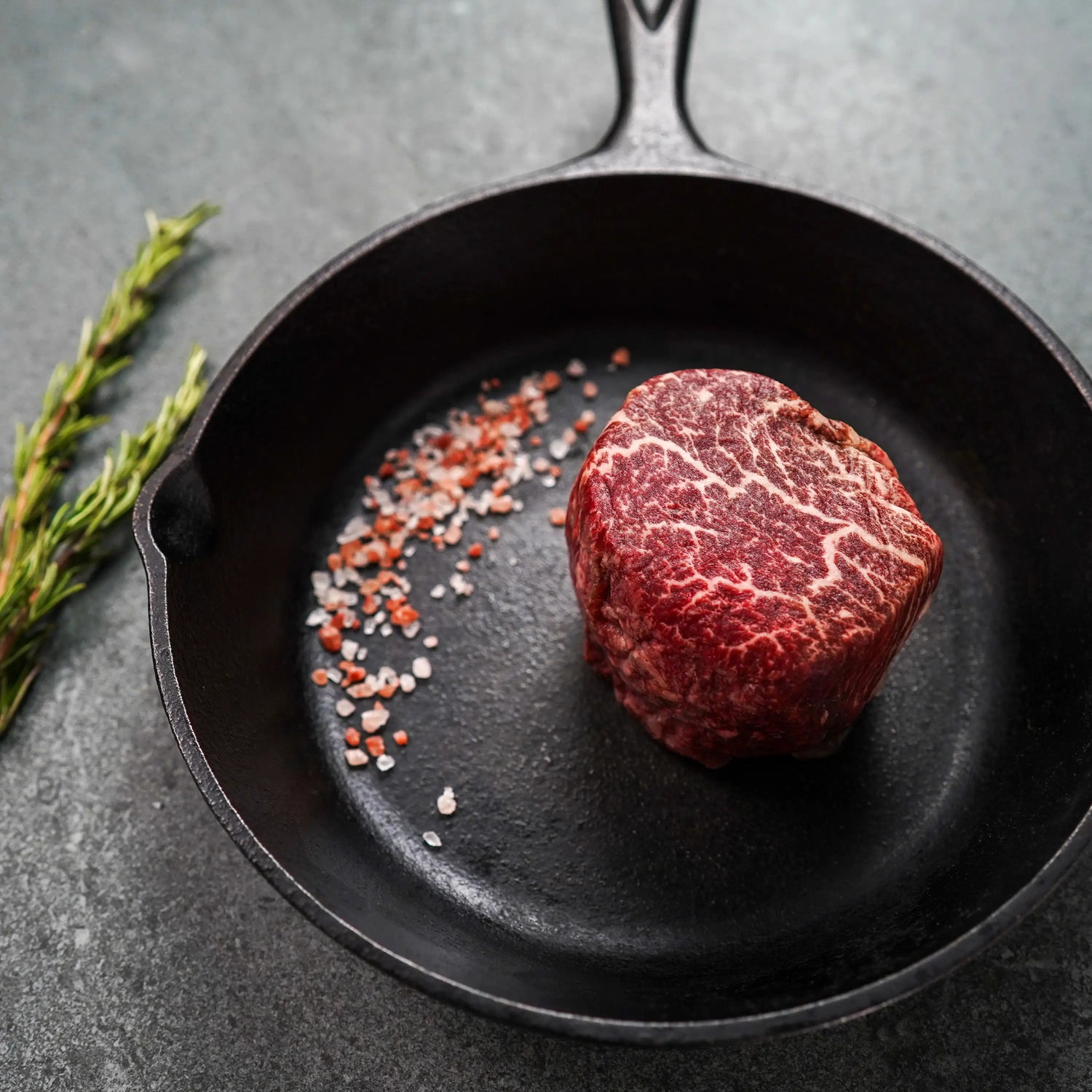 raw steak in iron pan with pink salt garnish and rosemary on dark surface