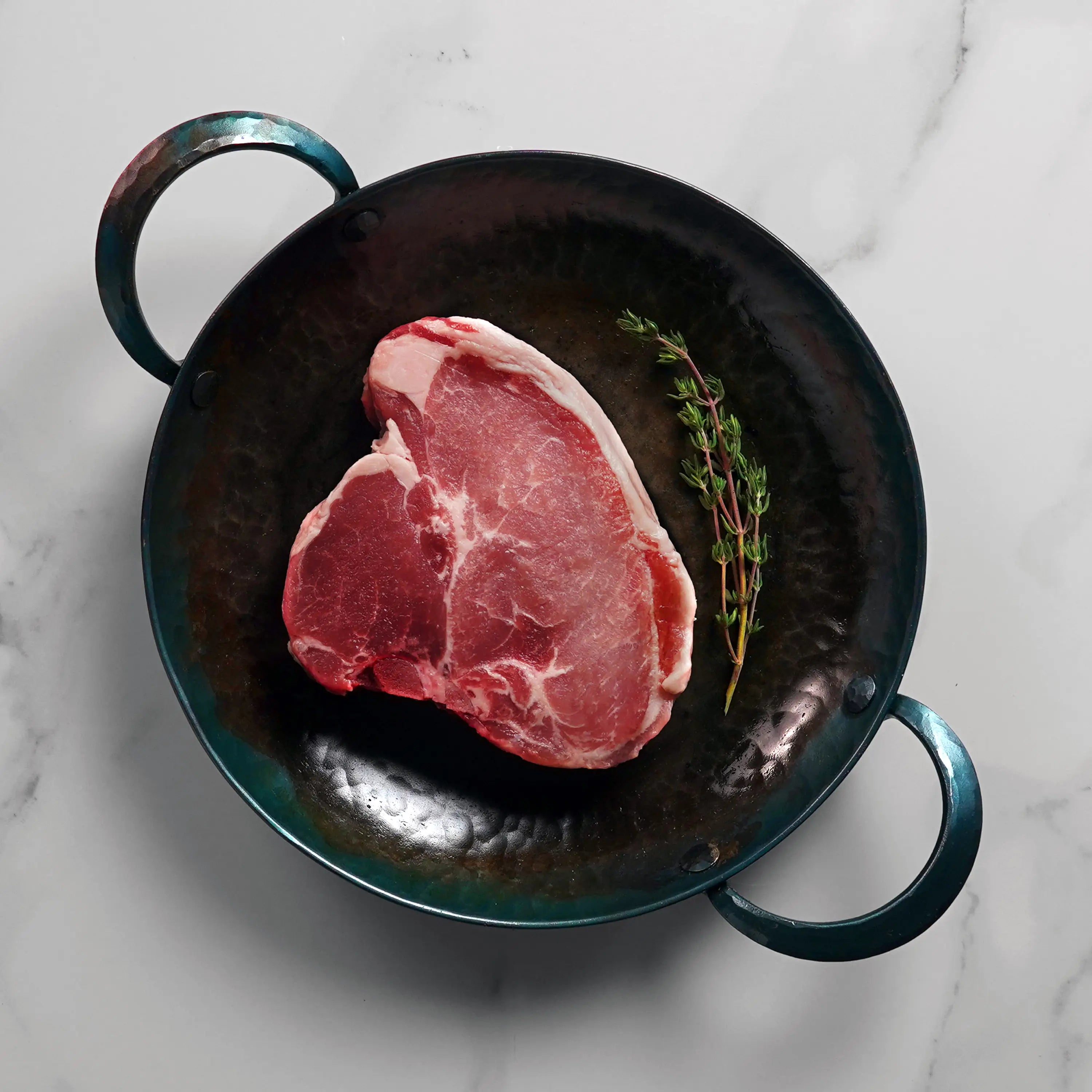 Raw pork chop on dark pan with herb garnish on marble stone