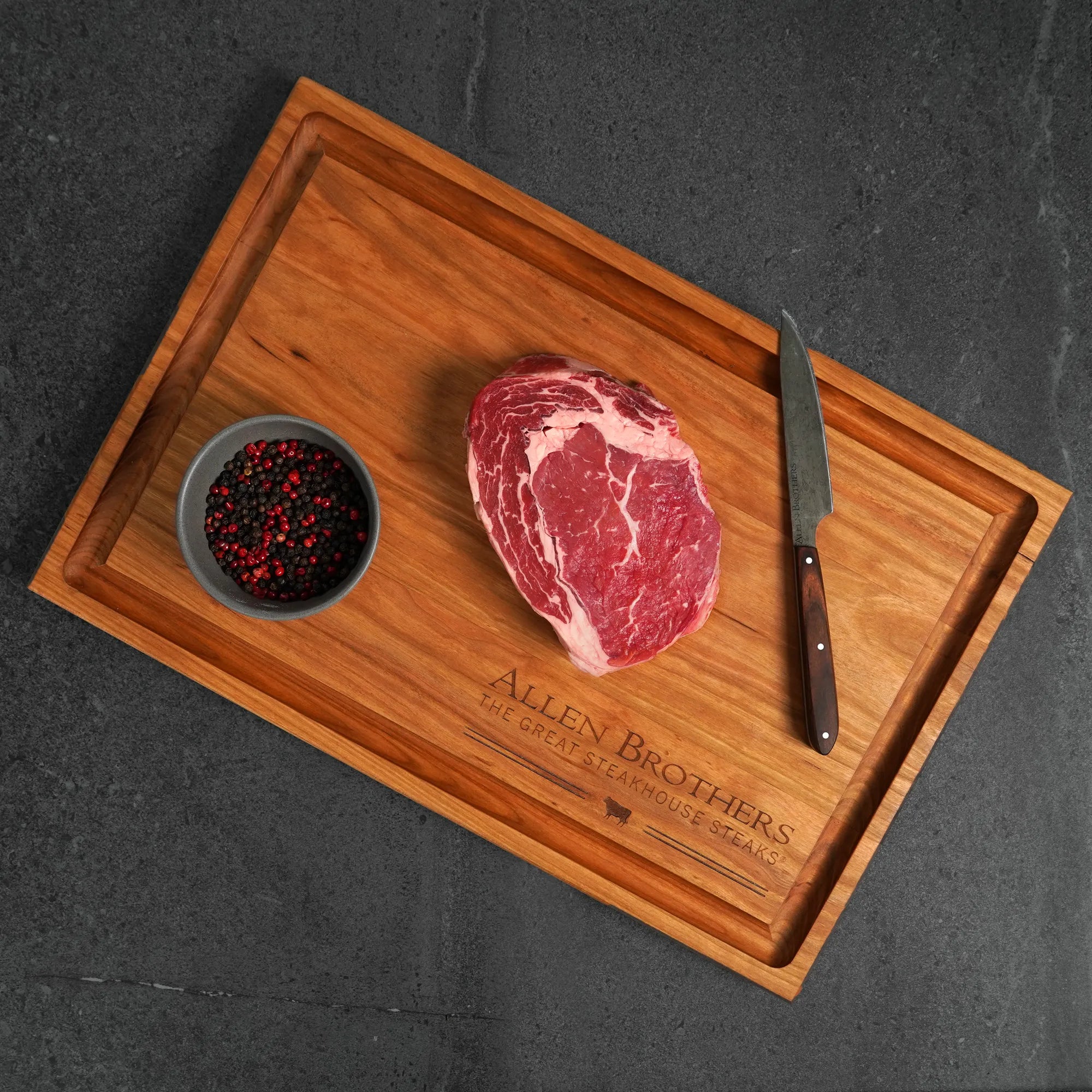 Wooden cutting board with a raw steak, small bowl of spices, and knife on a dark surface.