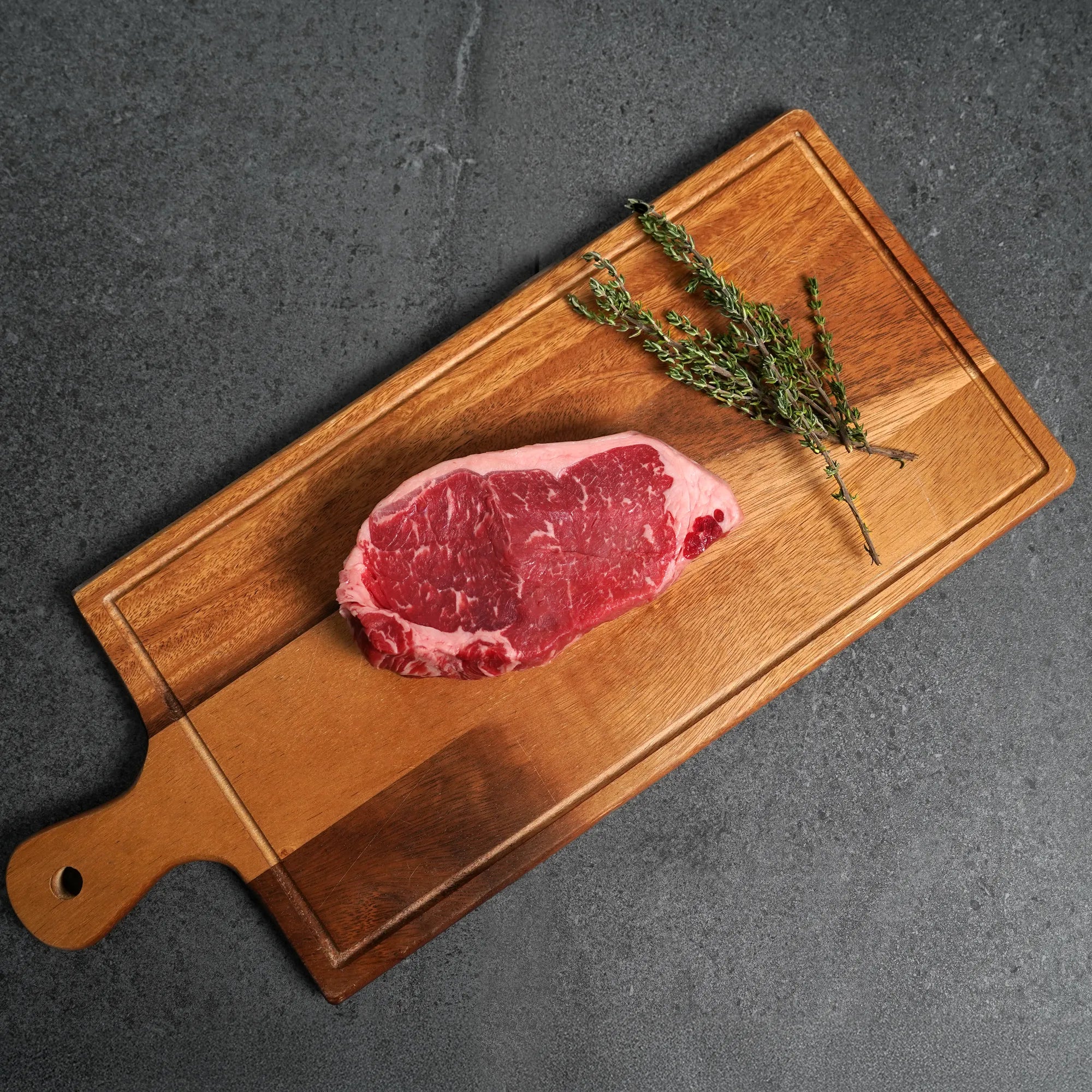 Raw steak on a wooden cutting board with a sprig of thyme against a dark background