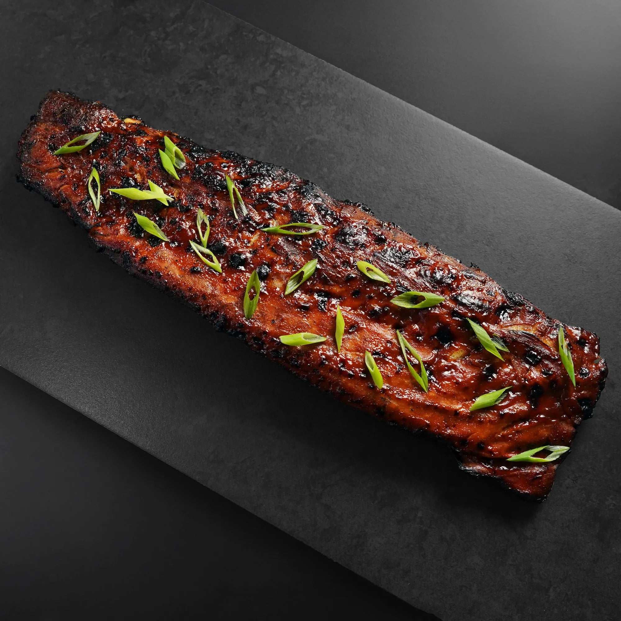 Heritage Duroc Pork Peeled Baby Back Rib, richly glazed and charred, garnished with scallions, presented on a dark platter, showcasing premium barbecue appeal.