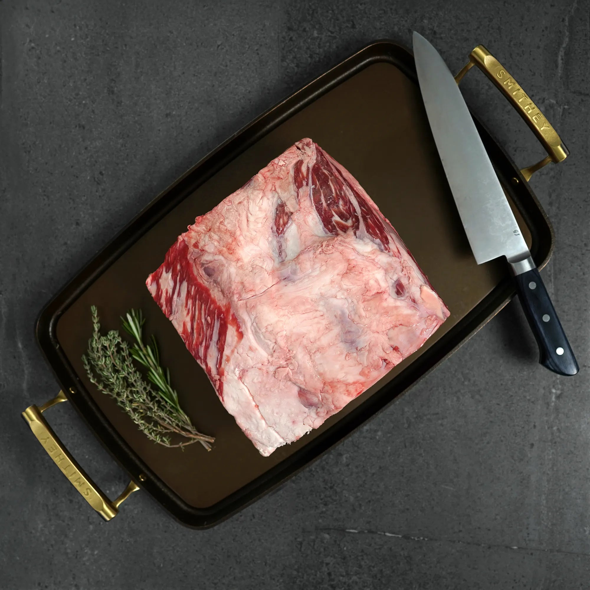 Raw meat on a dark tray with a knife and herbs on a gray background