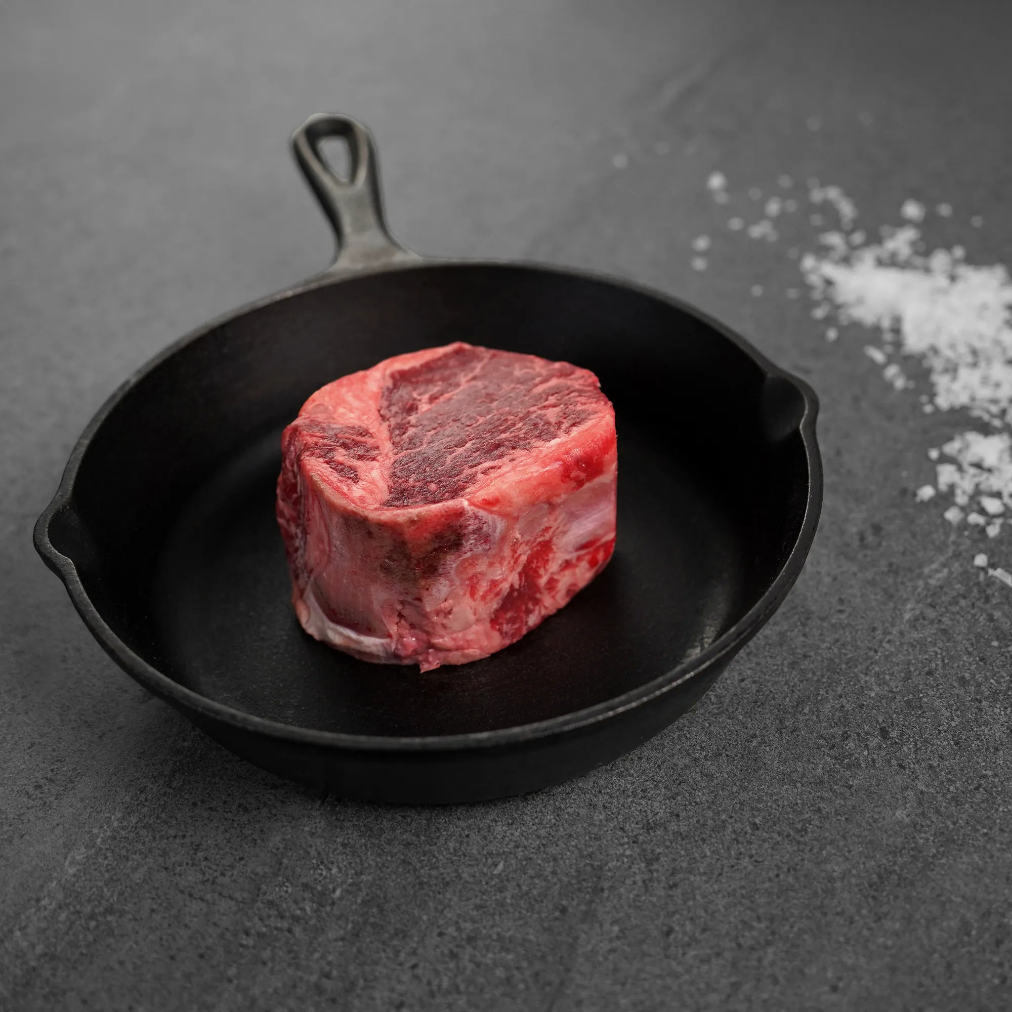 Raw steak in a black cast iron skillet on a dark surface with salt crystals.