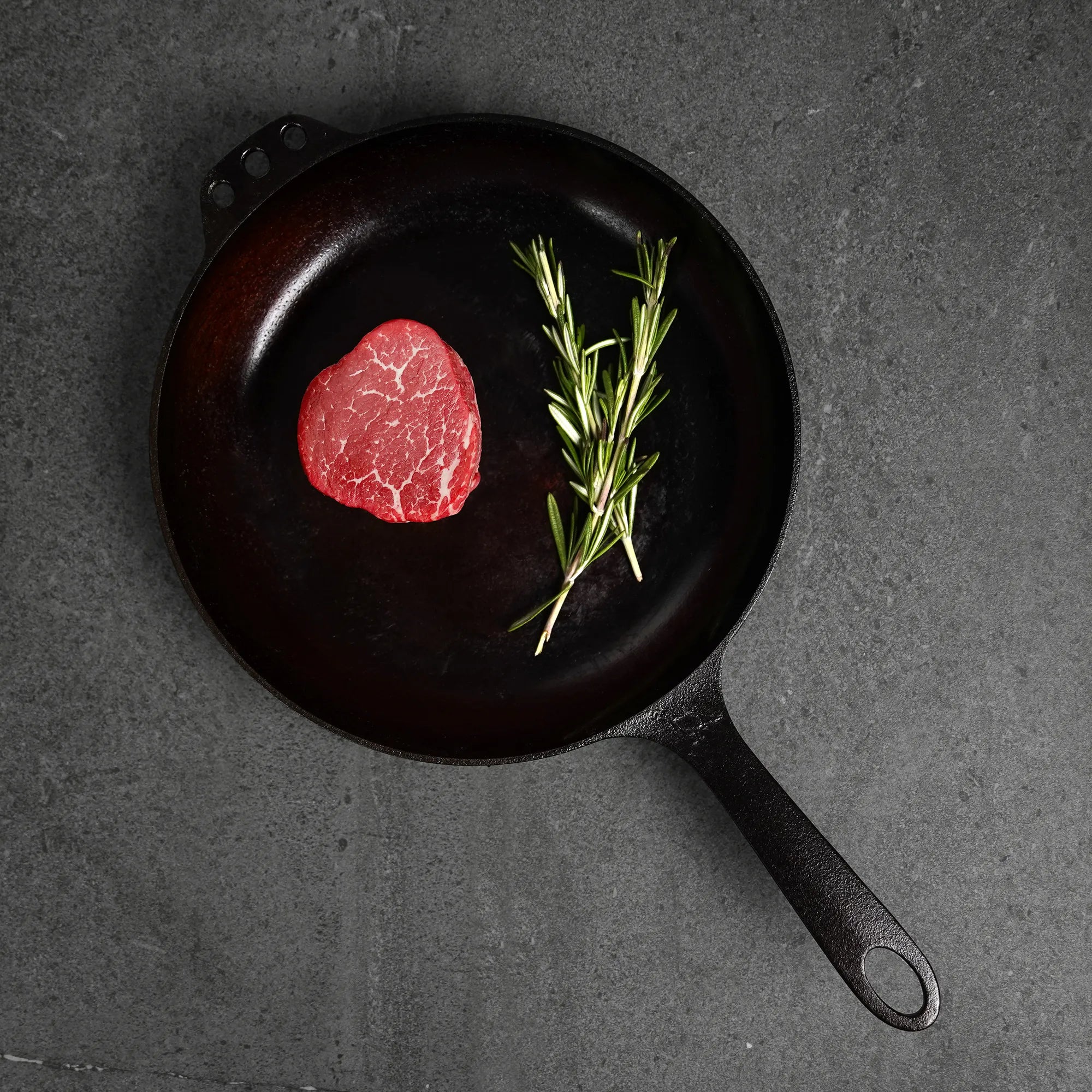 Raw steak with rosemary on a black cast iron skillet against a dark background