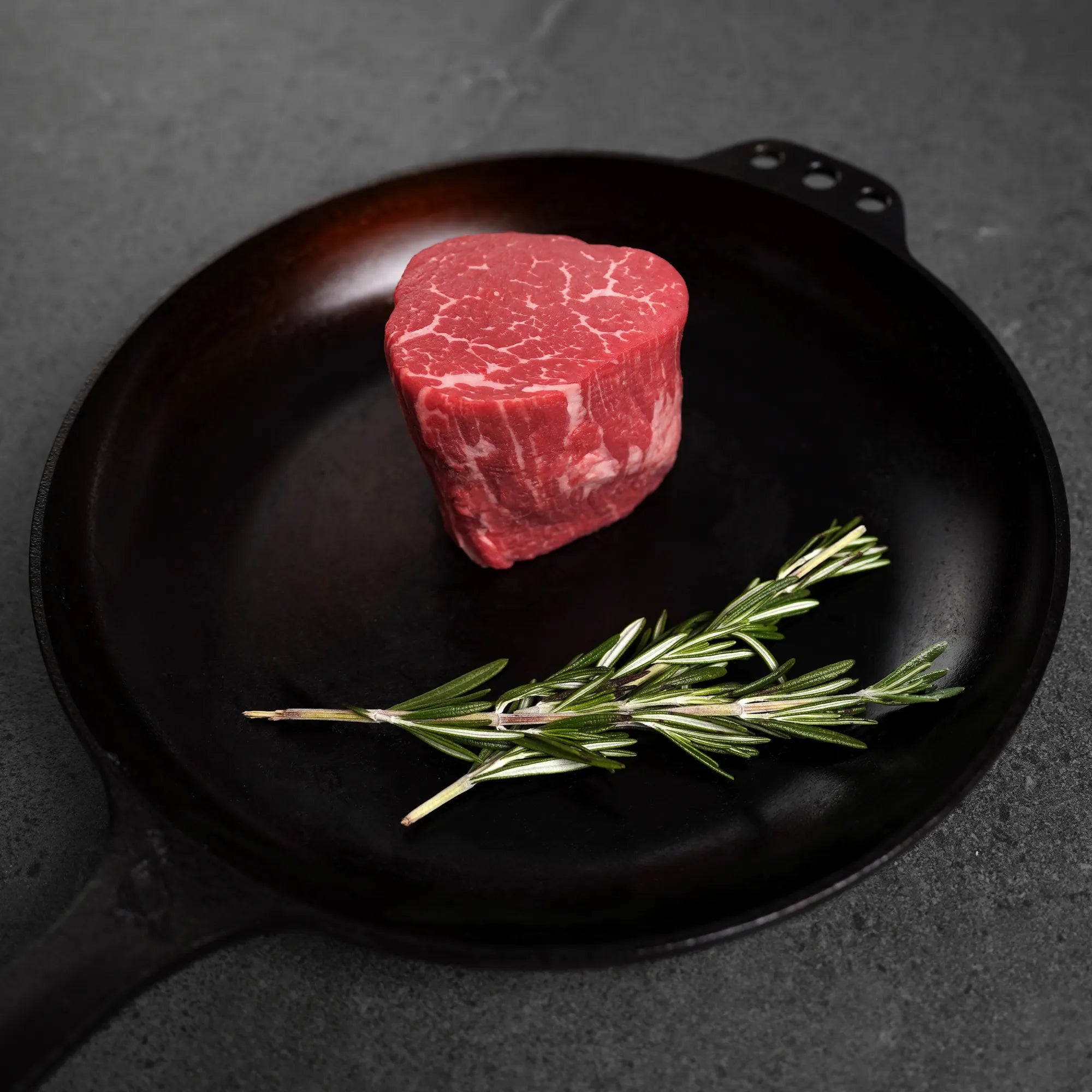 Raw steak on black iron pan with herbs on dark surface.