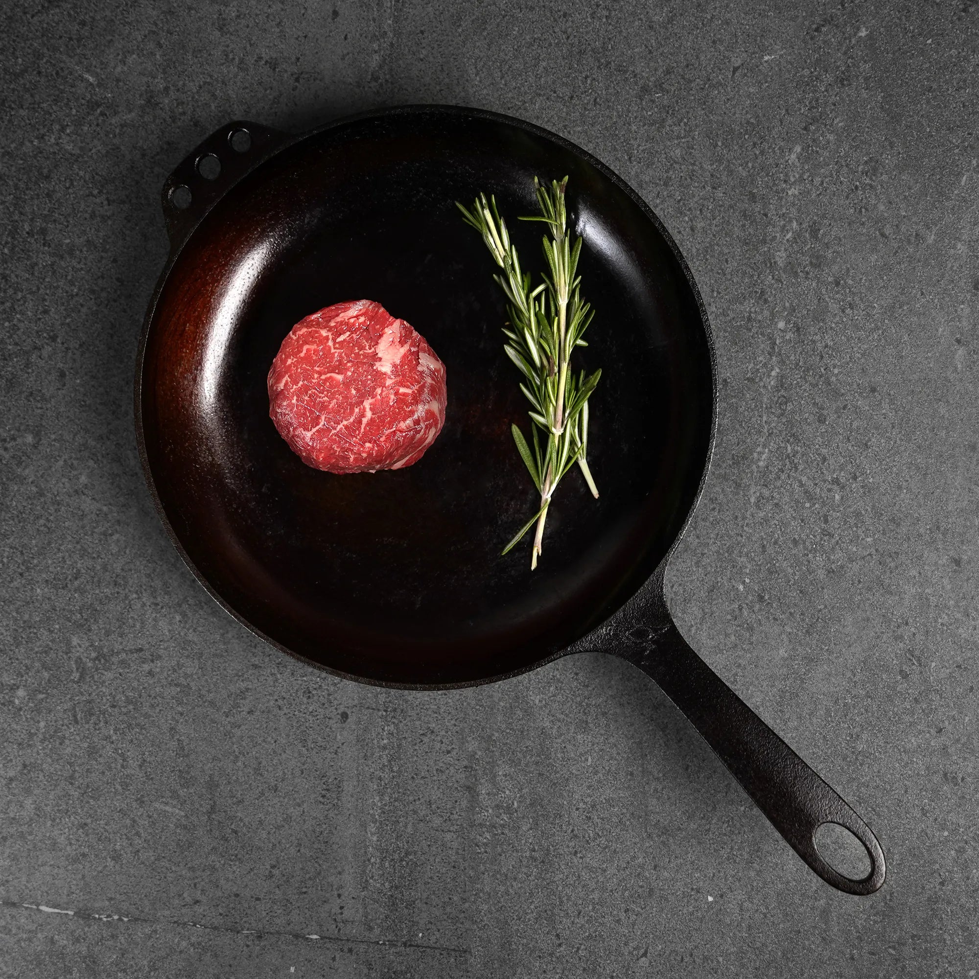Raw steak on black iron pan with herbs on dark surface.