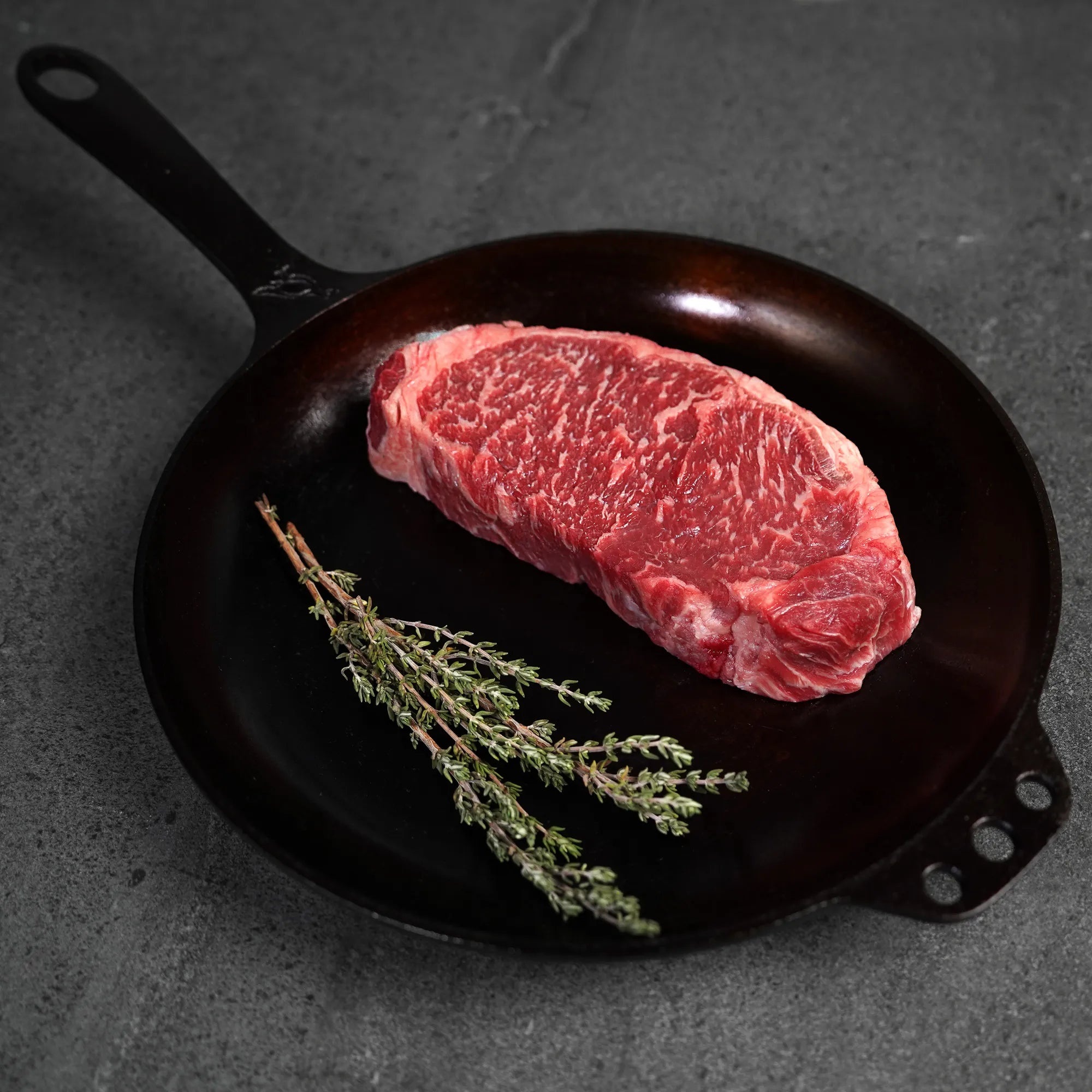 Raw steak on a black cast iron skillet with fresh herbs on a dark surface