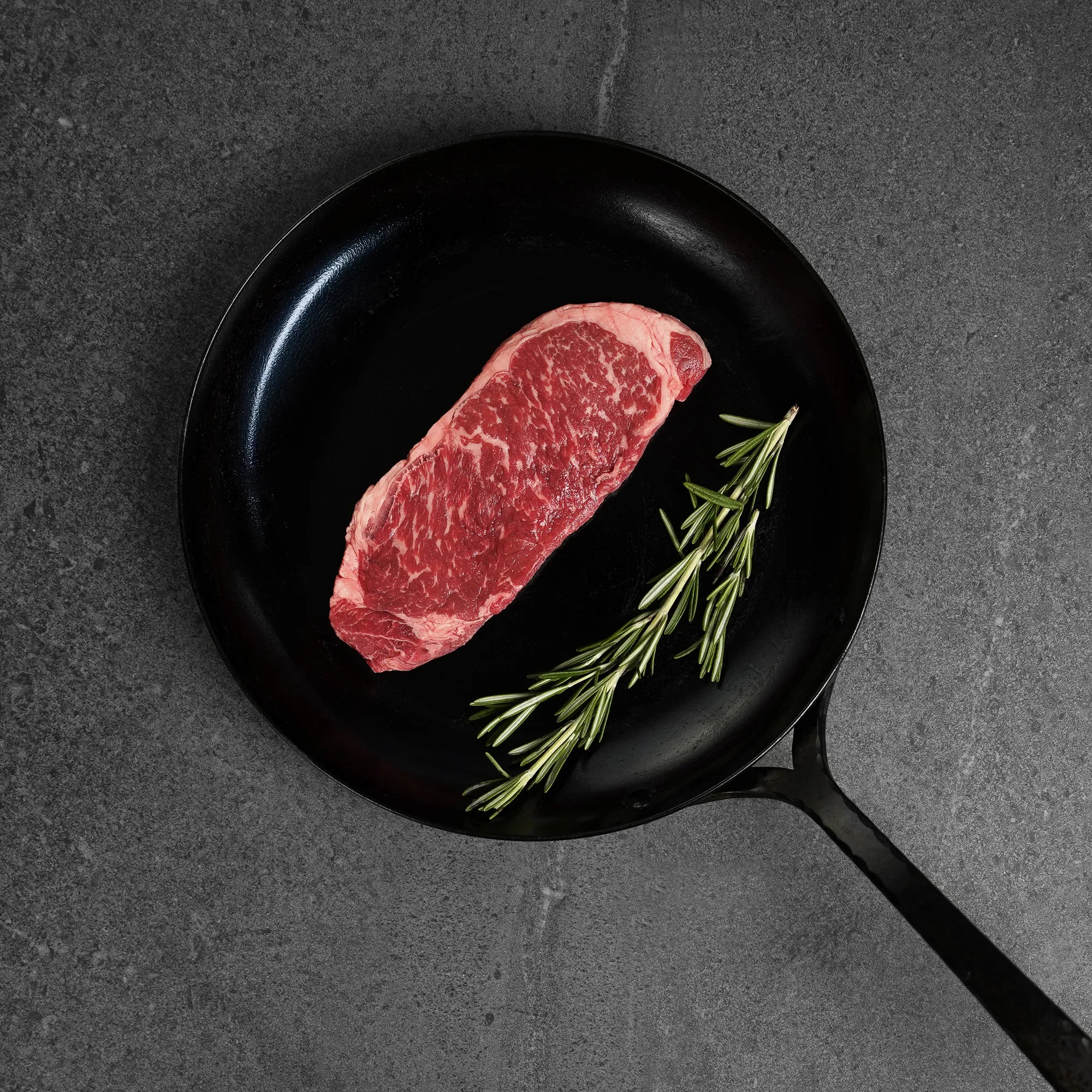 Raw strip steak on black iron pan with herb