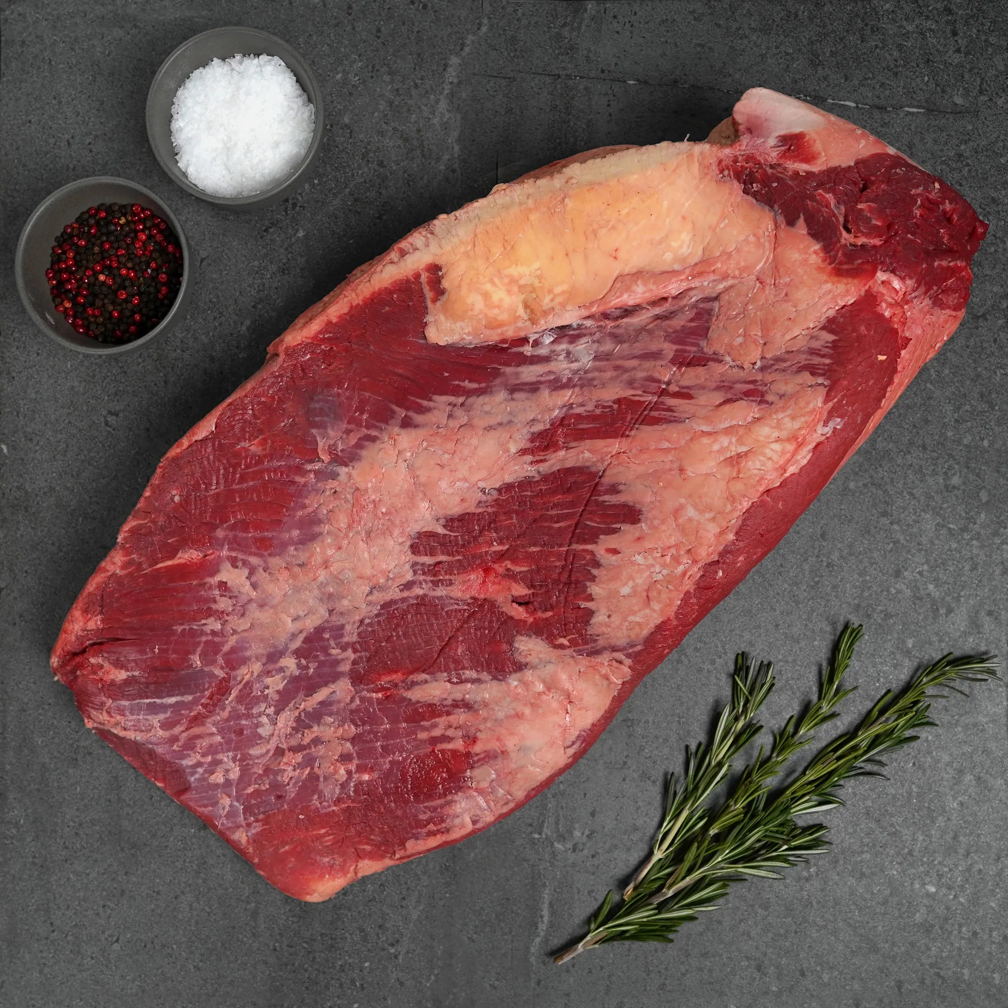 Raw meat with salt, pepper, and rosemary on a dark surface