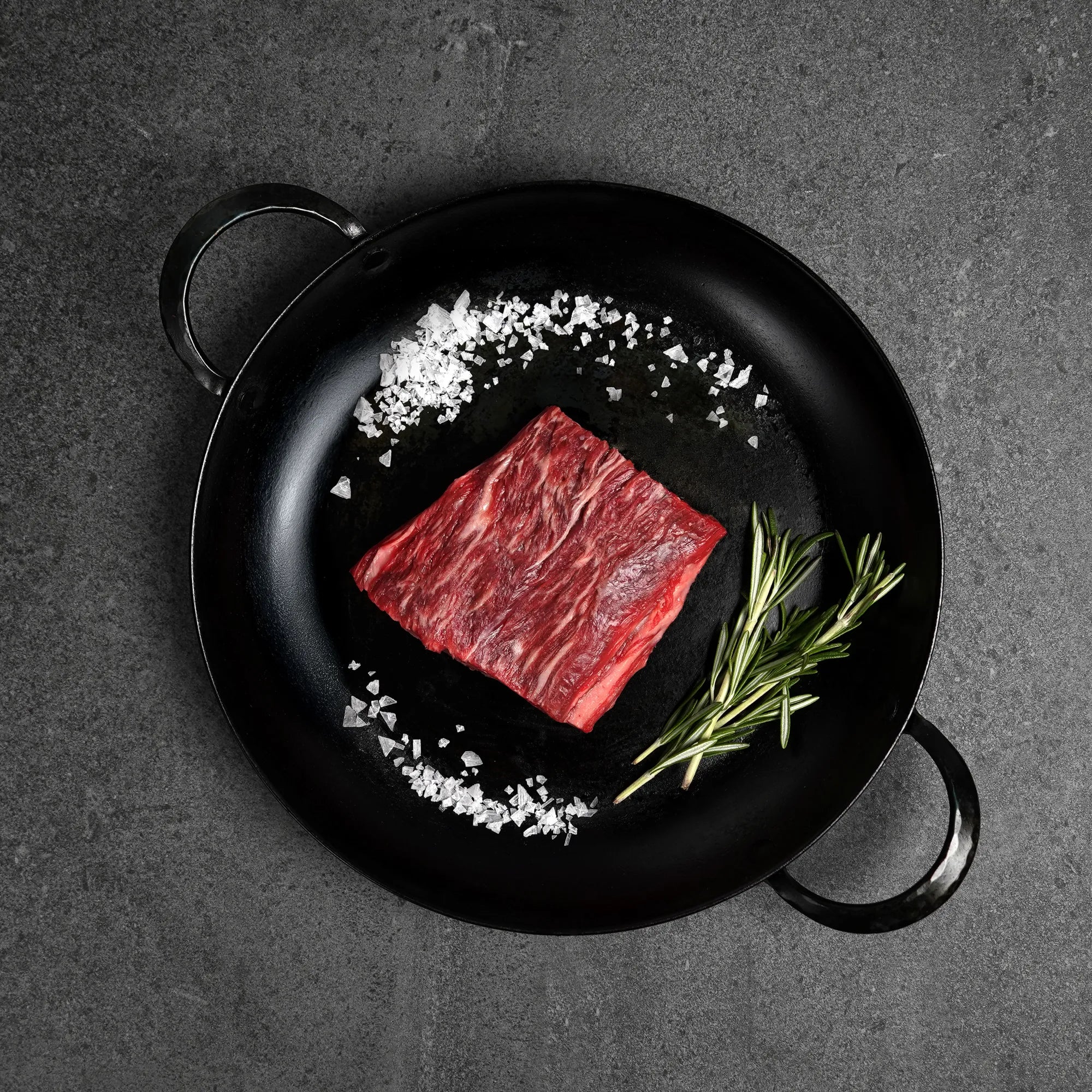 Raw steak with salt and rosemary on a black pan against a dark background
