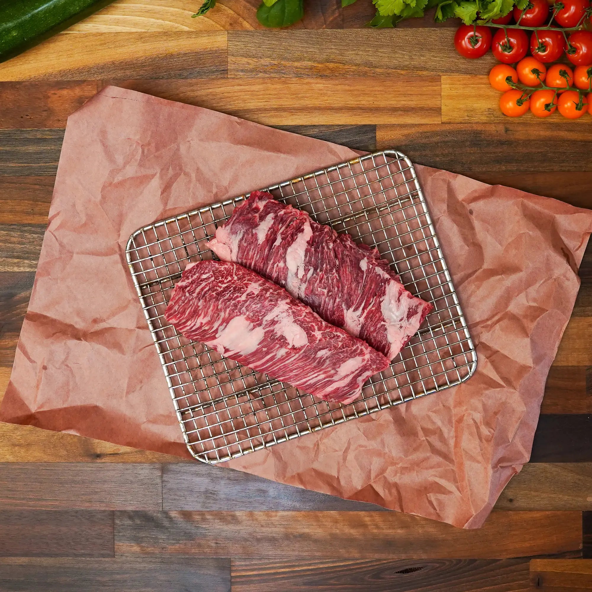 Prime Skirt Steak on a wire rack, showcasing its well-marbled texture, with cherry tomatoes, herbs, and cucumber visible on a wooden surface.