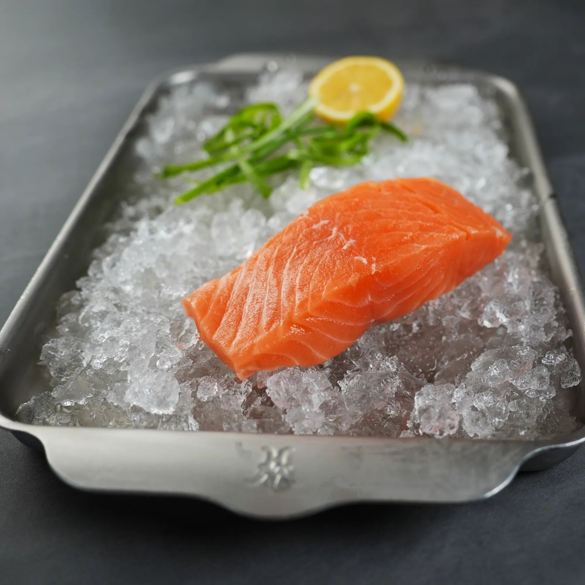 Salmon fillet on ice with a lemon wedge and greenery in a metal tray.