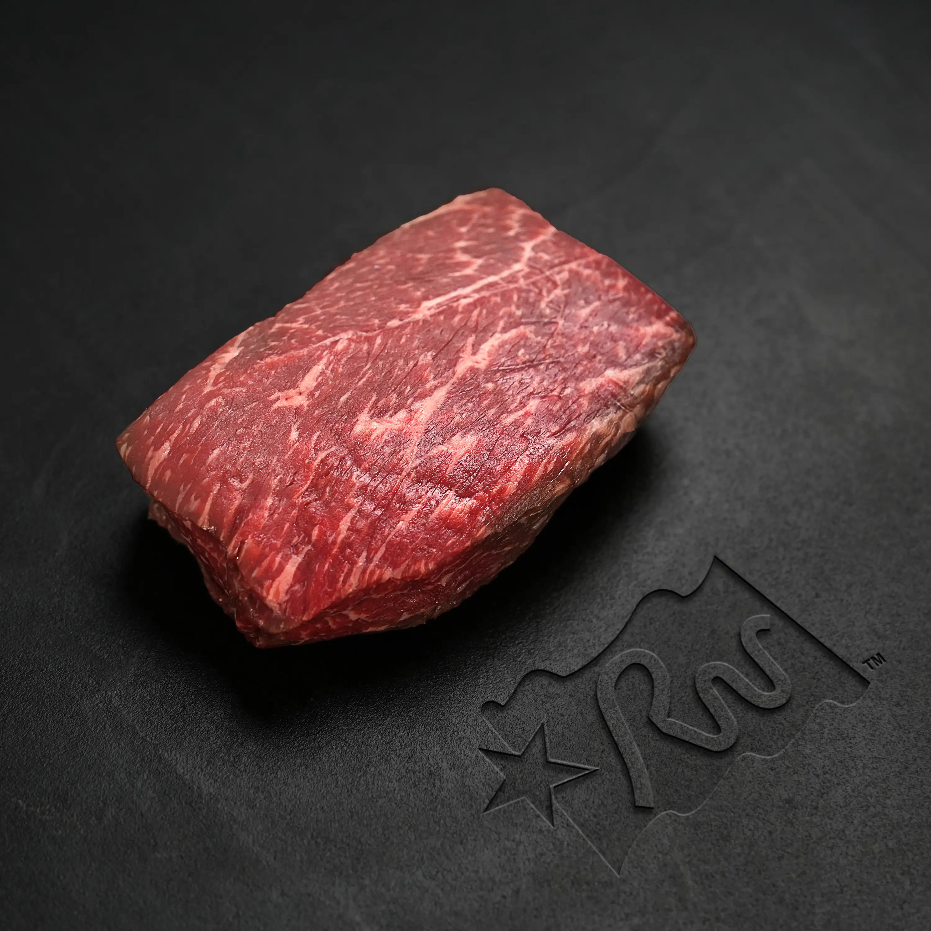 Texas Wagyu Sirloin Bavette Steak, a thick-cut marbled piece, rests on a dark surface, showcasing its tender texture ideal for high-heat cooking.