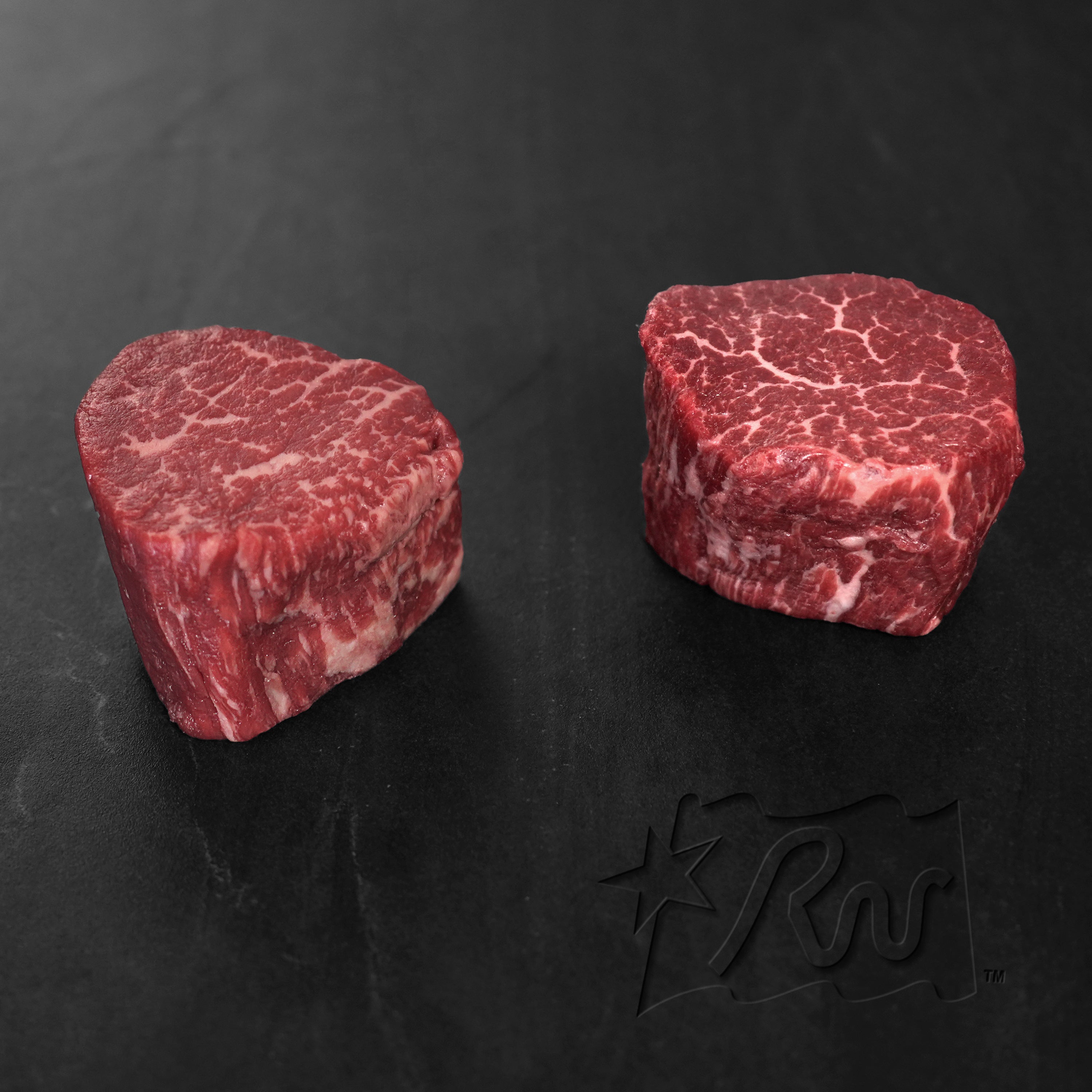 Two pieces of raw beef on a dark surface with a brand logo.