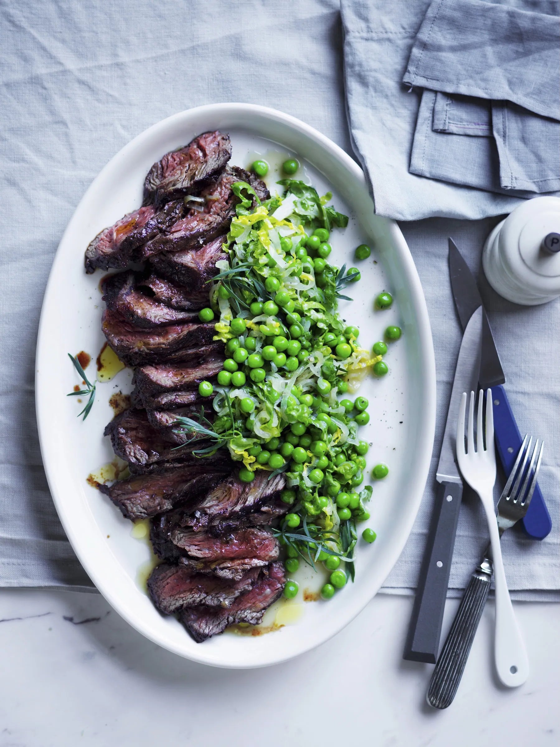 Plated dish of sliced meat with green peas on a white plate, set on a gray surface with cutlery.