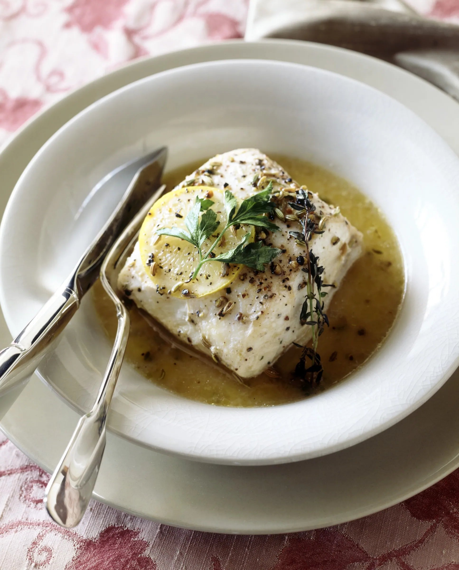 White plate with a piece of fish garnished with herbs, served with a lemon wedge and a knife on a patterned tablecloth.